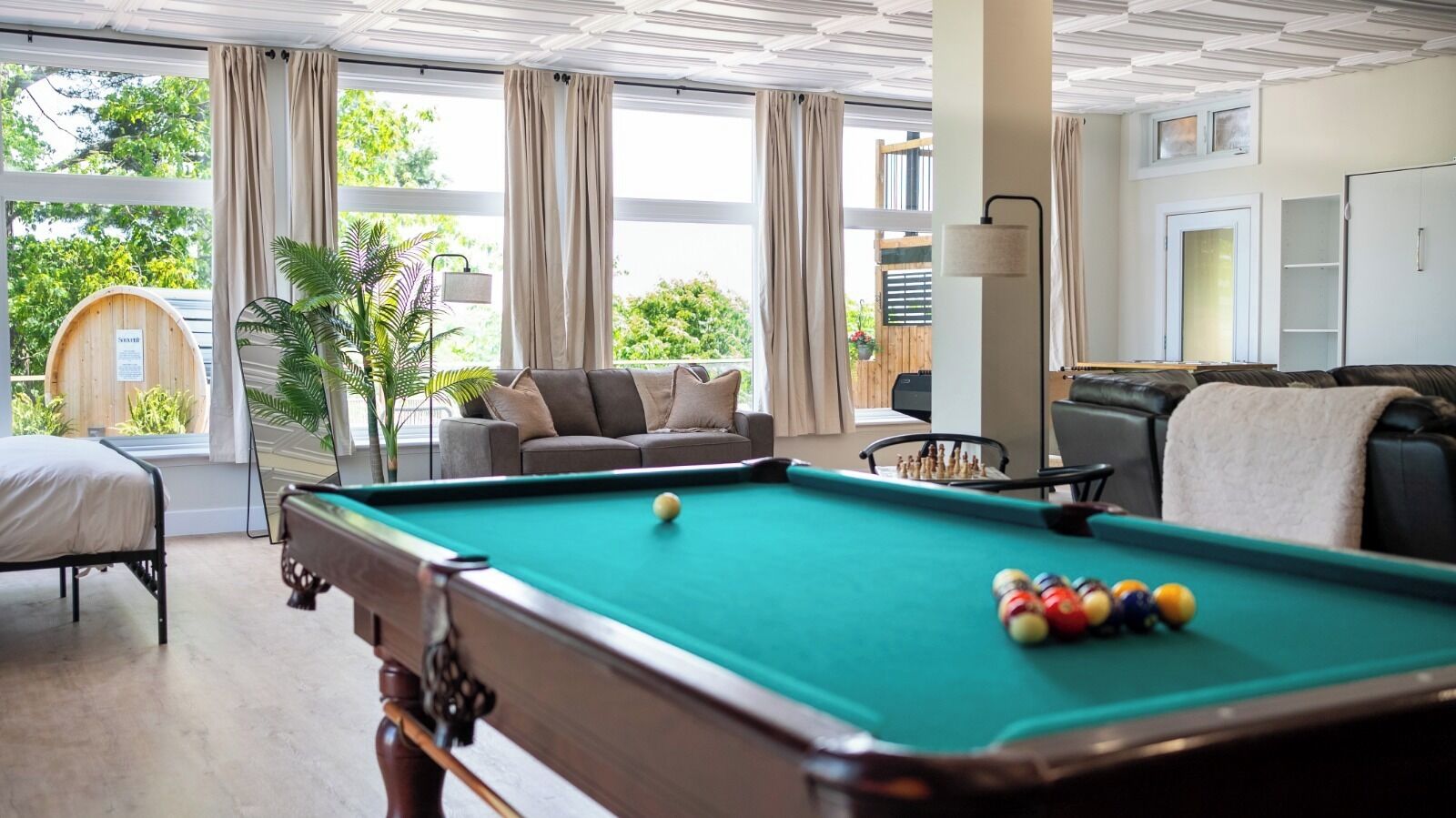 Una mesa de billar con bolas, un sofá y una silla en una habitación con ventanas amplias y cortinas.