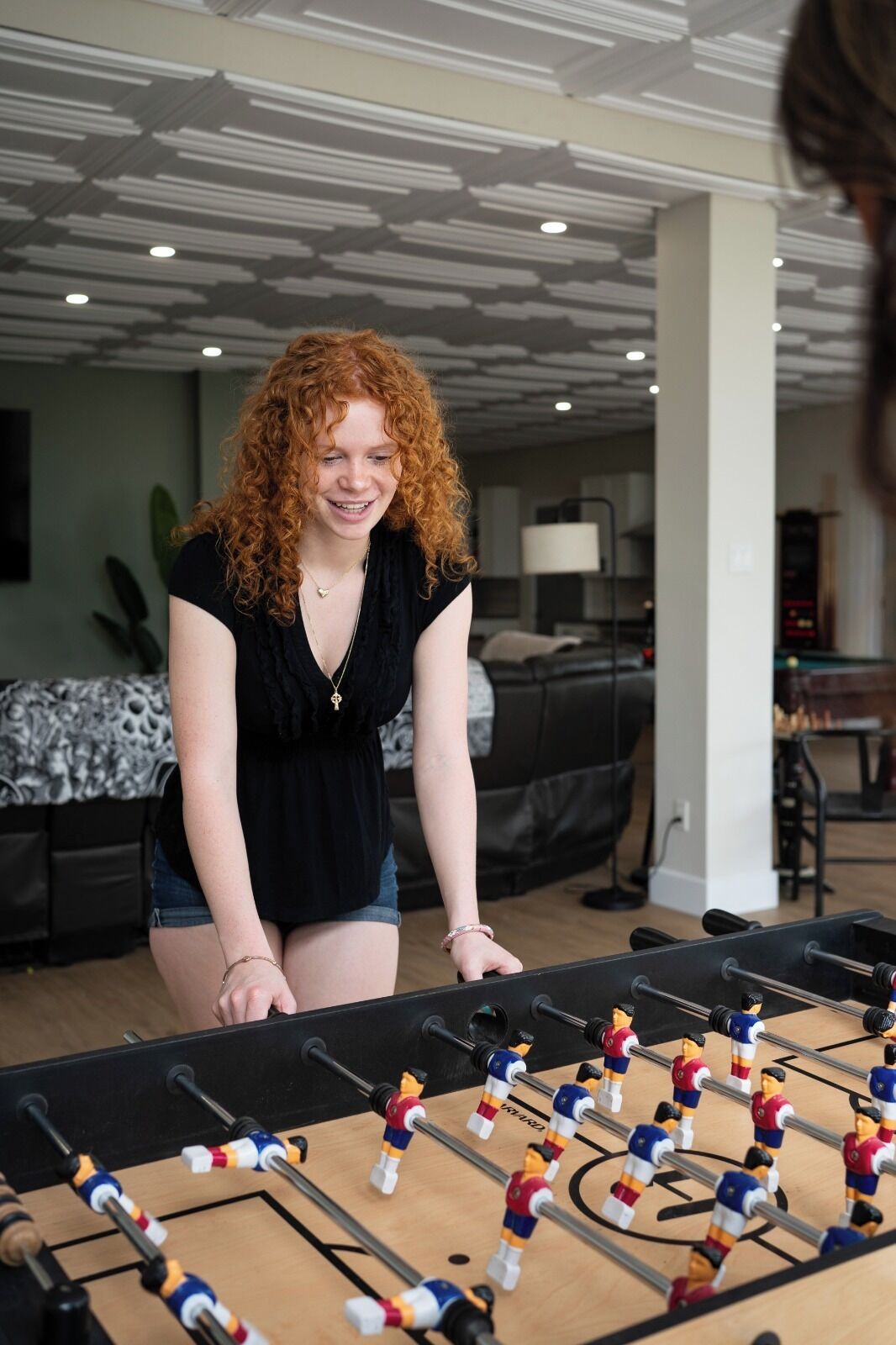 A person playing foosball in a room with a bed and a lamp in the background.