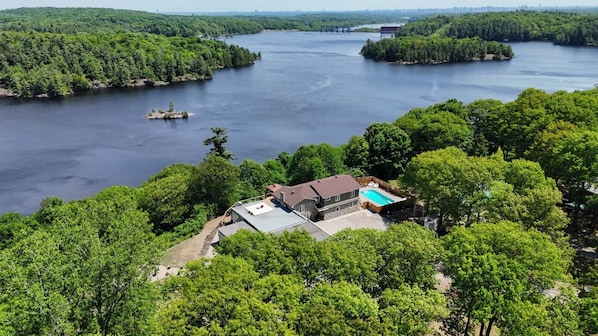 A house with a swimming pool, surrounded by dense forest and a large body of water.