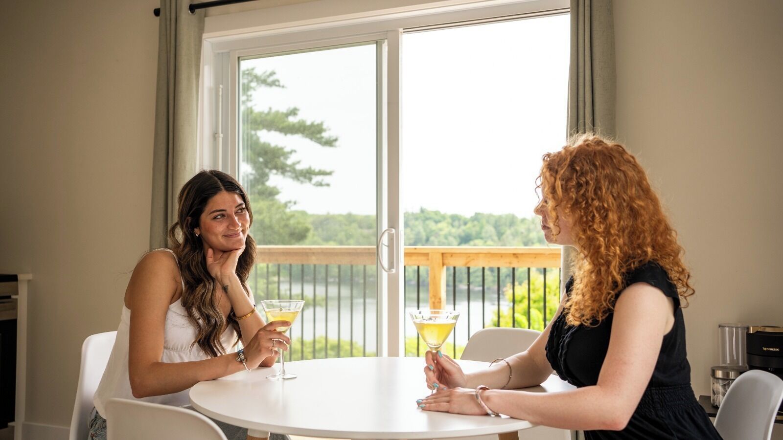 Dos personas en una mesa con bebidas, vistas a follaje y una masa de agua a través de una ventana.
