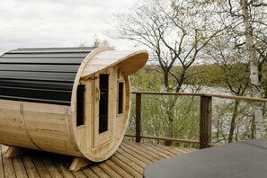Una sauna de madera con techo curvo, ubicada en una plataforma de madera con vista a árboles y un cuerpo de agua.