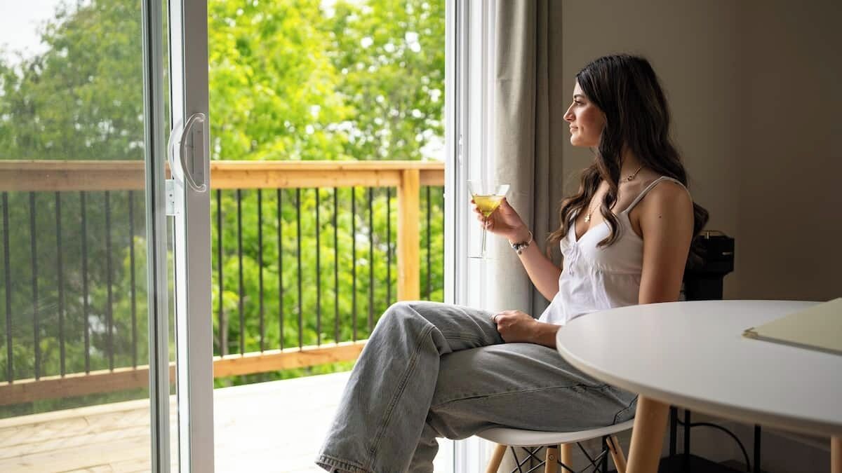 Une femme est assise près d’une fenêtre, d’où l’on aperçoit une terrasse en bois et de la verdure à l’extérieur.