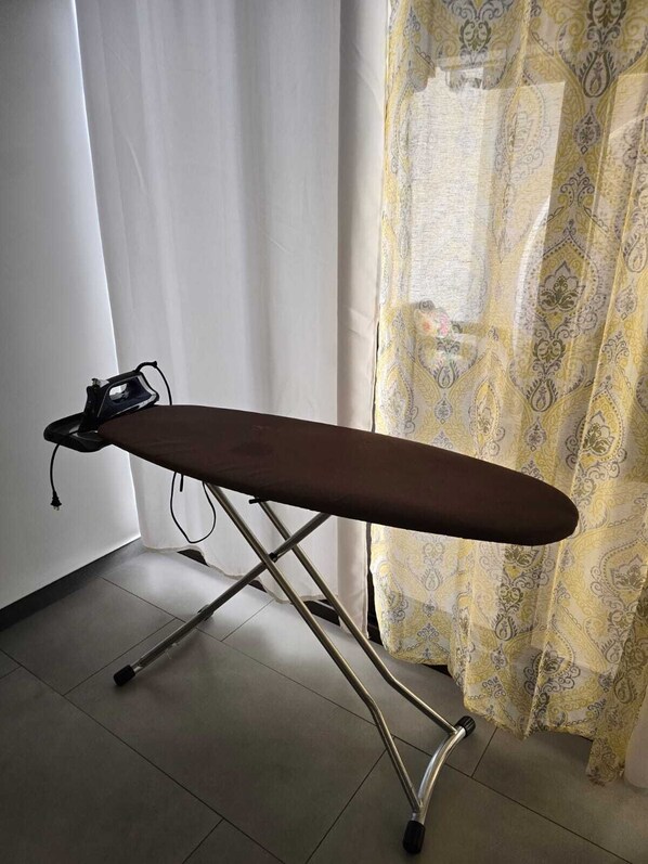 An ironing board with a power cord, positioned in a room with a curtain and a window.