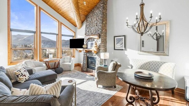 A spacious living room with a stone fireplace, large windows, and a chandelier. - Traverse Hospitality (Crested Butte)