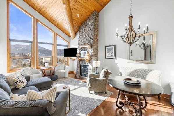 A spacious living room with a stone fireplace, large windows, and a chandelier.