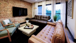 A living room with a brick wall, a flat-screen TV, a brown leather sofa, a green armchair, and a wooden coffee table.