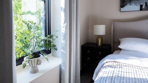 A bedroom with a bed, a bedside table with a lamp, a window with curtains, and a potted plant on the windowsill.
