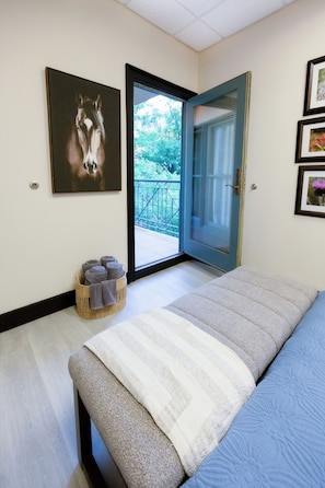 Un dormitorio con cama, puerta que da a un balcón, un cuadro de caballos enmarco y una cesta con toallas.