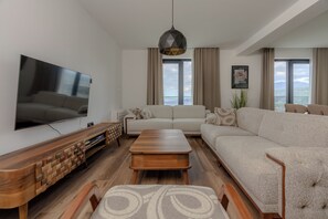 Ein modernes Wohnzimmer mit Flachbildfernseher, Couchtisch aus Holz, beiger Couch und großem Fenster mit Aussicht.