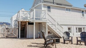 Een huis aan het strand met een balkon en een trap naar de begane grond, plus strandstoelen.