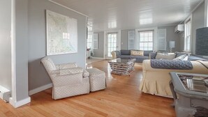 Een ruime woonkamer met een groot raam, een kaart aan de muur en een bank met een kussen waarop 'BEACH' staat.