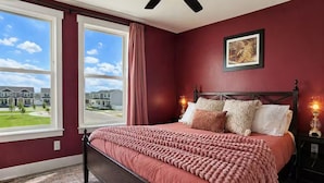 A bedroom with a large window, a bed with a pink blanket, and a view of houses outside.