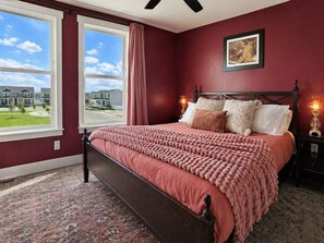 A bedroom with a large window, a bed with a pink blanket, and a view of houses outside. - Moss & Blush-King Bed-Garage (Billings)