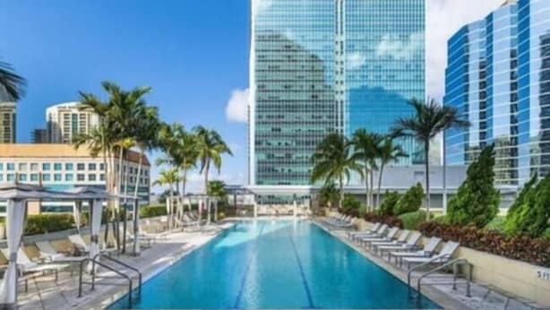 A swimming pool with lounge chairs, palm trees, and modern buildings in the background.