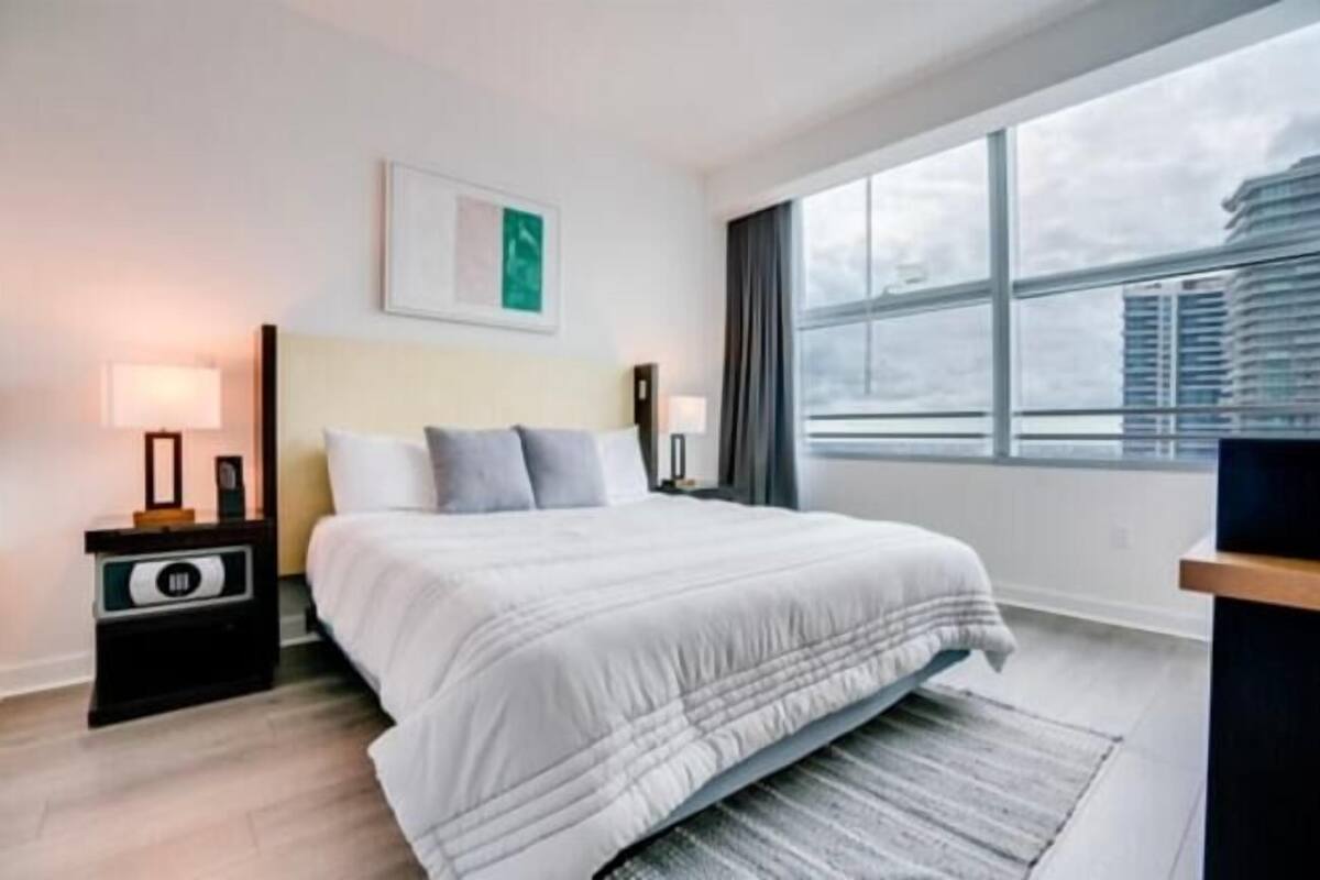 A hotel room with a bed, bedside lamps, a nightstand, a window with a view of buildings, and a framed picture on the wall.