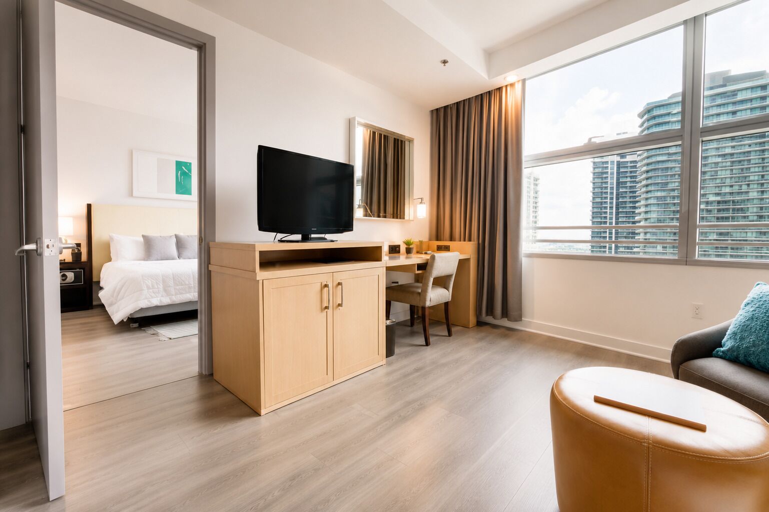 A modern hotel room with a bed, a desk with a TV, a chair, and a large window with a city view.