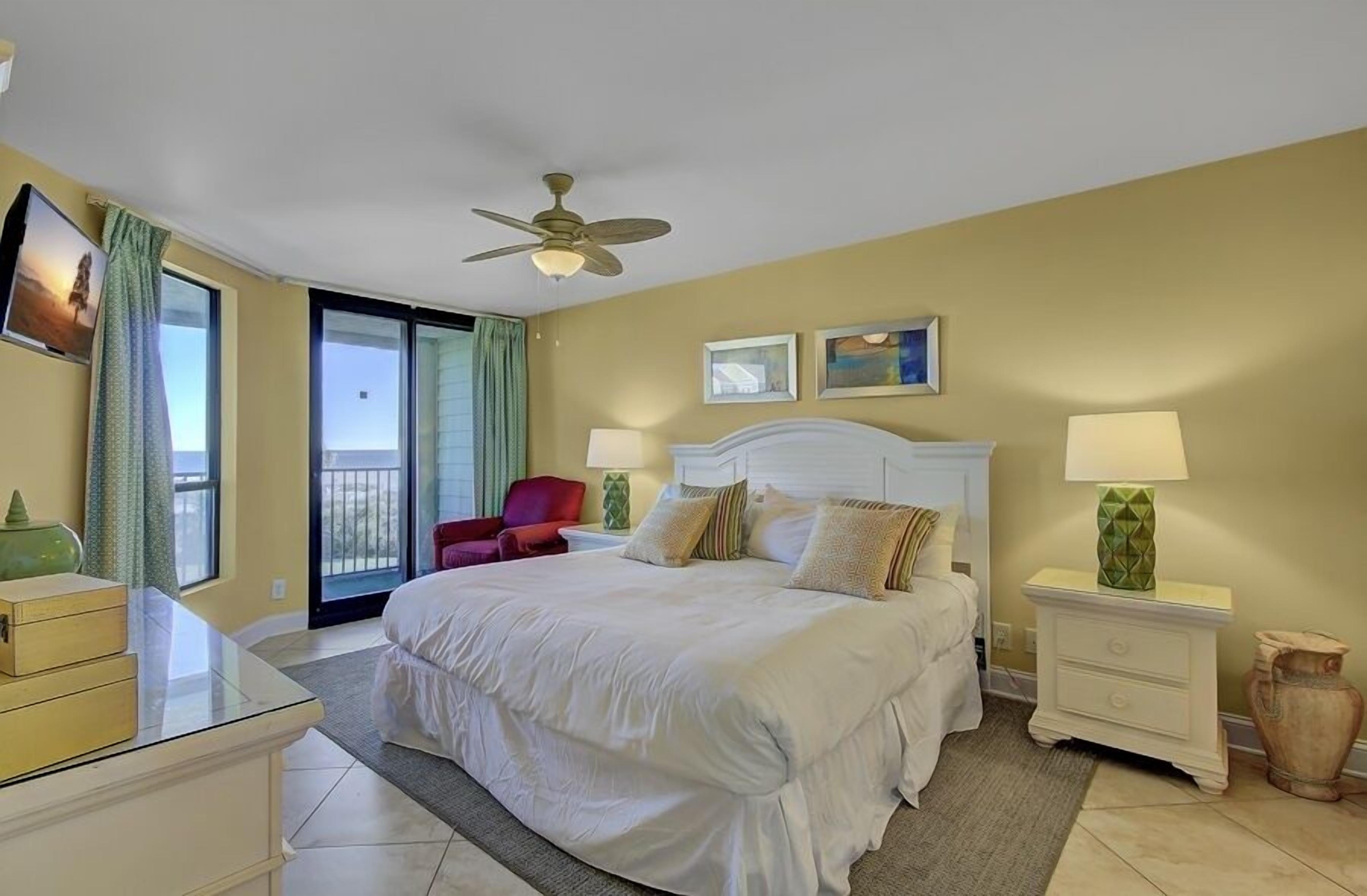 A hotel room with a bed, two bedside tables, a ceiling fan, a red chair, and a balcony with a view.