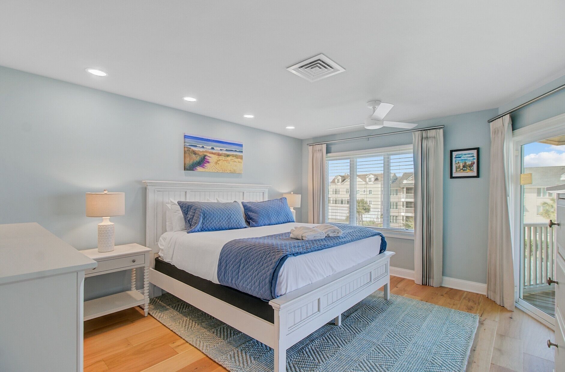 A bedroom with a bed, bedside tables, a ceiling fan, a window with curtains, and a view of buildings outside.