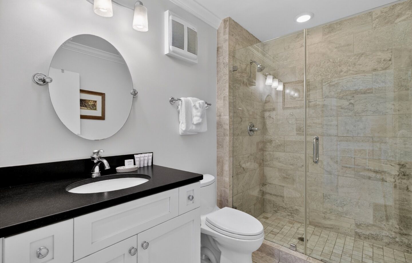 A modern bathroom with a round mirror, a black countertop sink, a toilet, and a glass-enclosed shower.