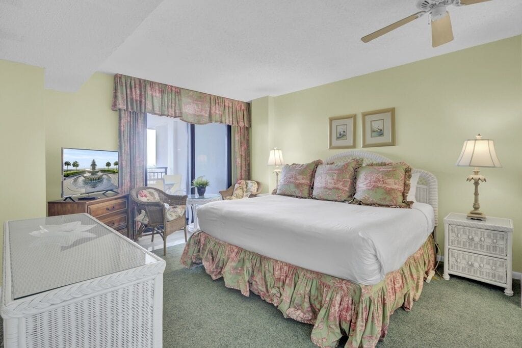 A bedroom with a bed, a desk, a chair, a television, and a ceiling fan.