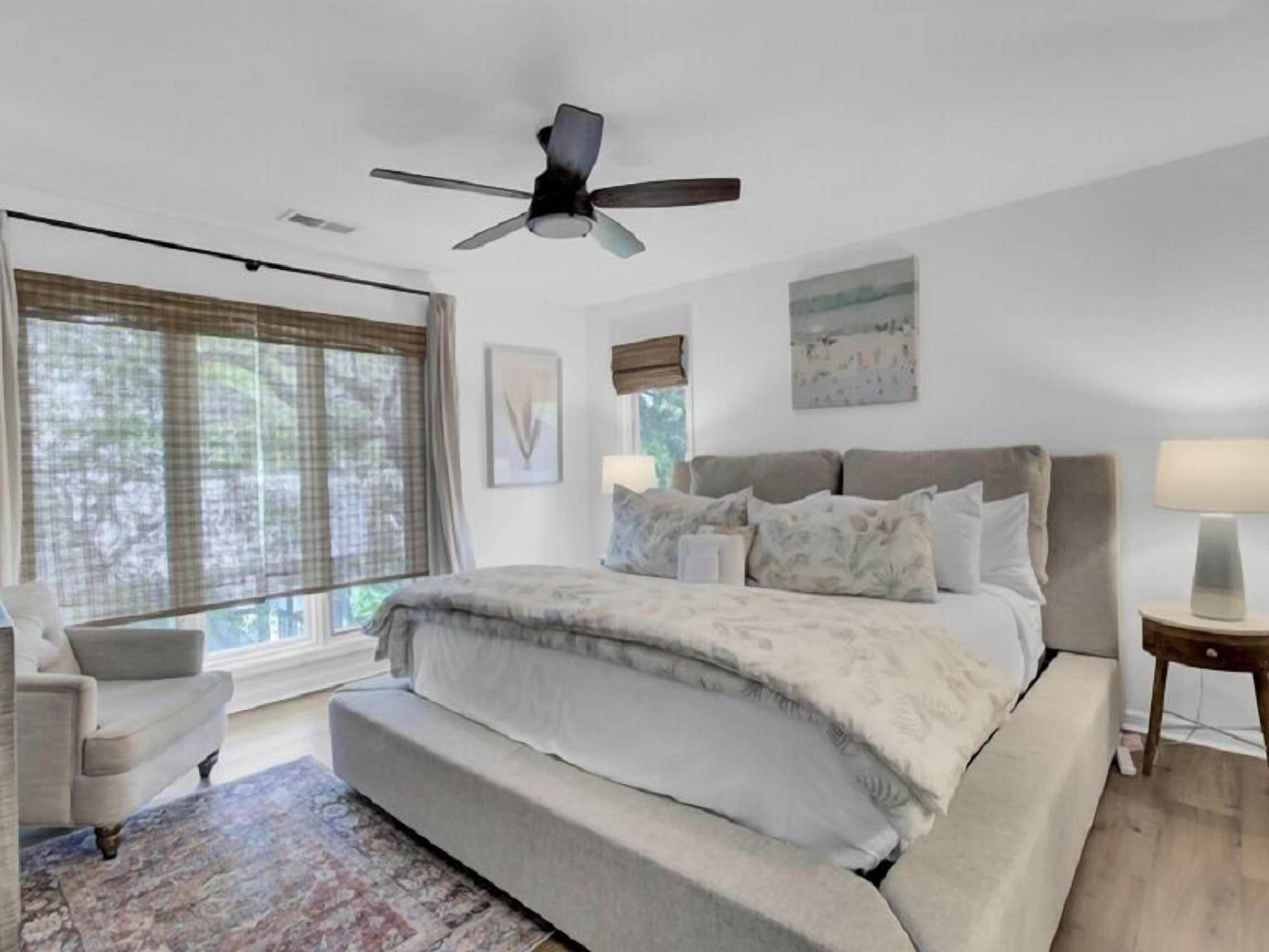 A bedroom with a large bed, a ceiling fan, a window with blinds, a chair, and a bedside table.
