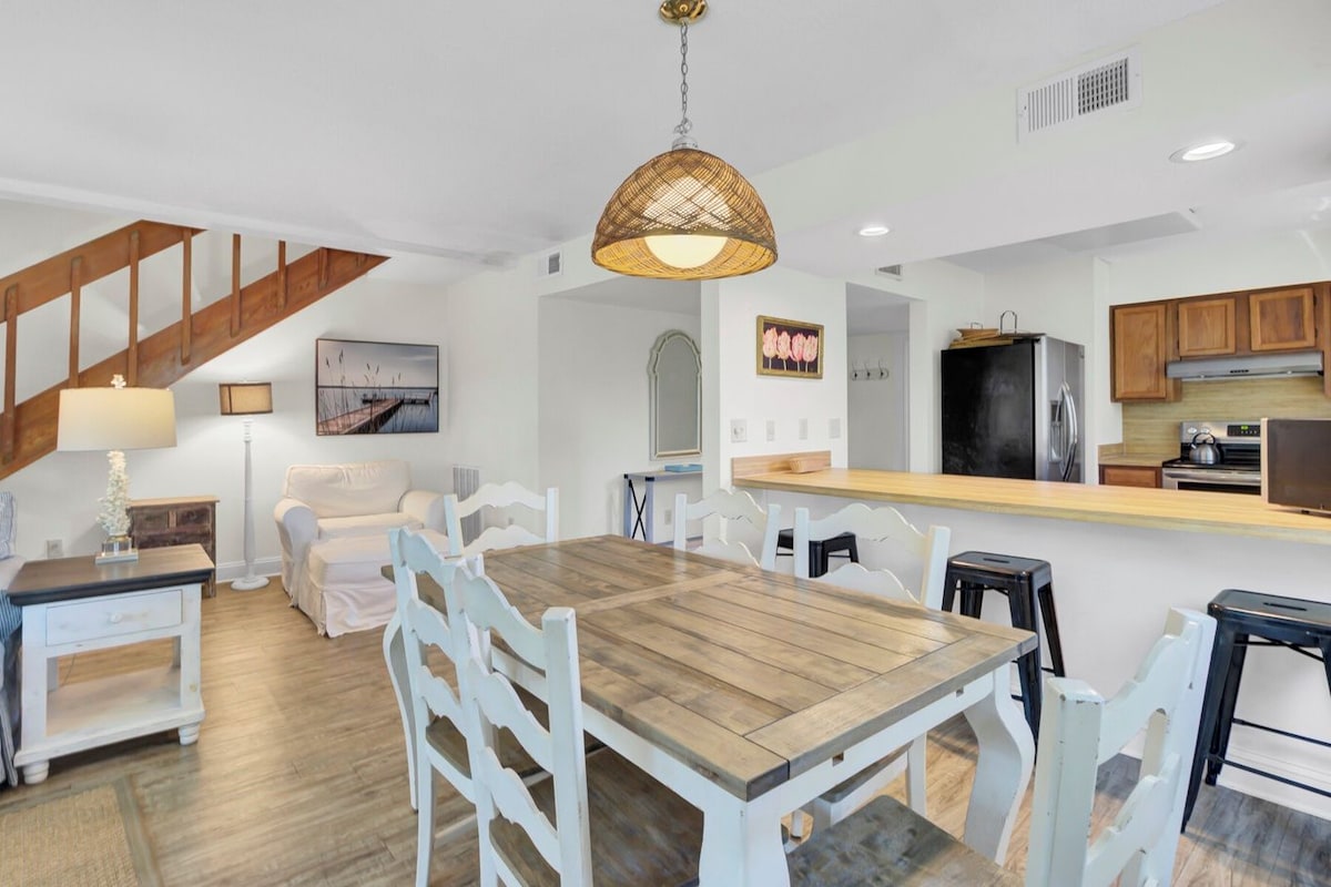 A modern kitchen with a wooden dining table and white chairs, a staircase with wooden railings, and a hanging light fixture.
