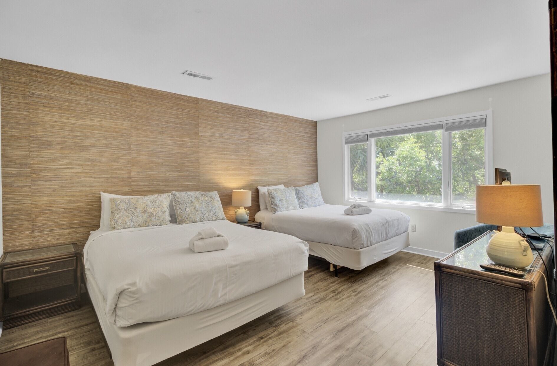 A hotel room with two beds, a wooden accent wall, a window with a view, and a desk with a lamp.