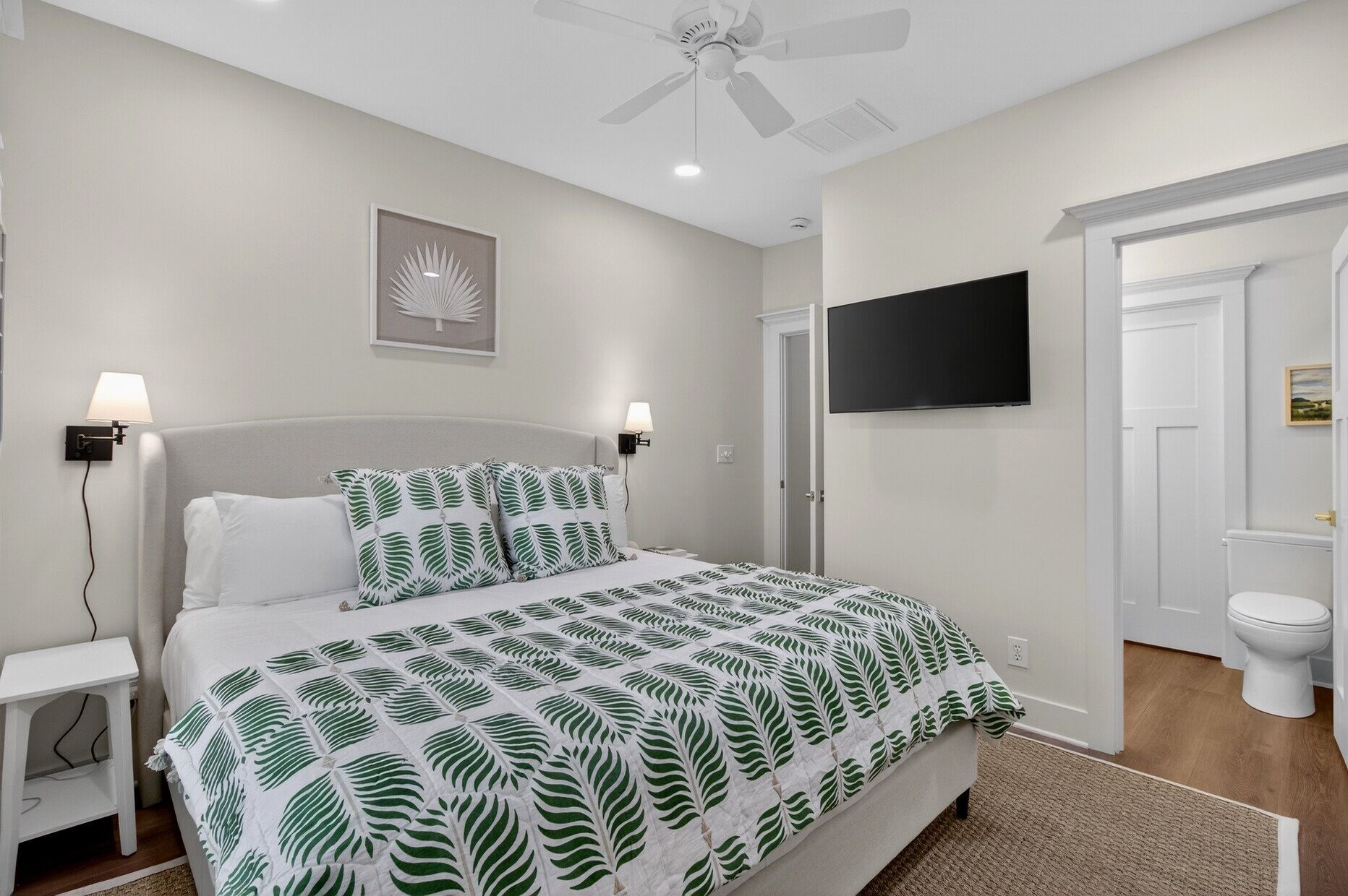 A bedroom with a bed, bedside lamps, a ceiling fan, a mounted TV, and a door leading to the bathroom.