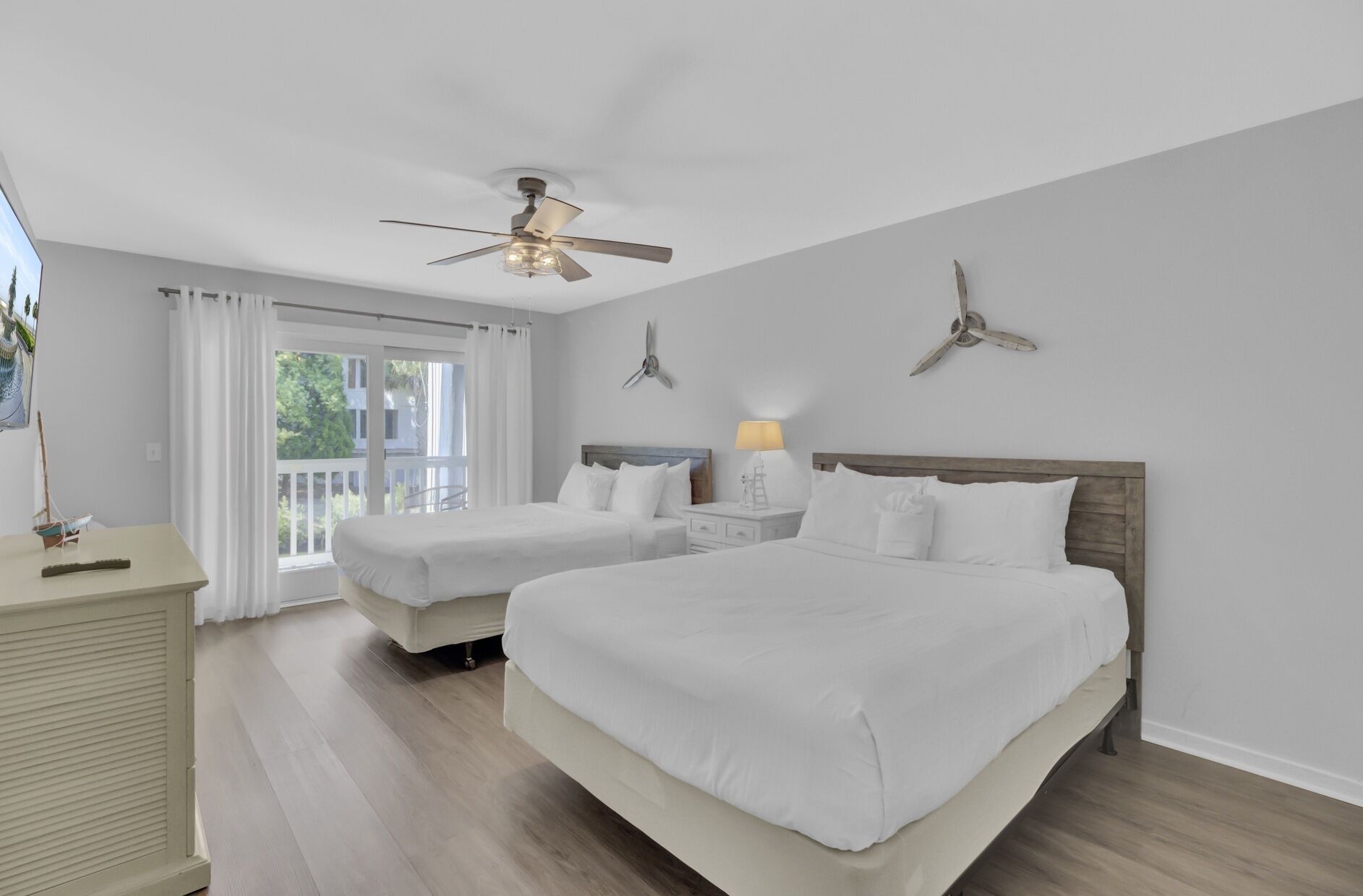 A hotel room with two beds, a ceiling fan, a nightstand with a lamp, and a view of the outdoors.