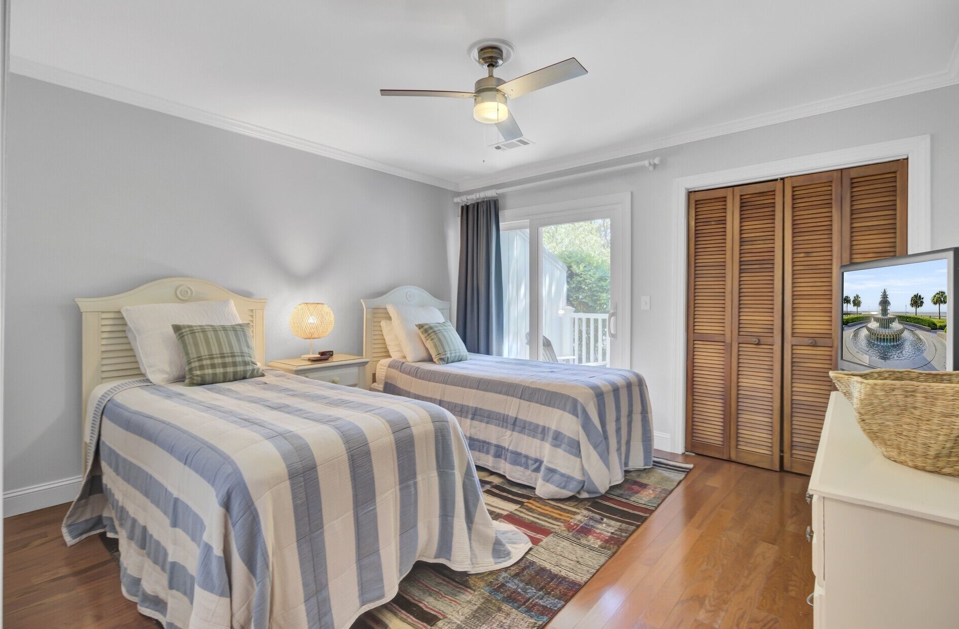 A bedroom with two beds, a ceiling fan, a nightstand with a lamp, a window with curtains, and a view of a water feature outside.