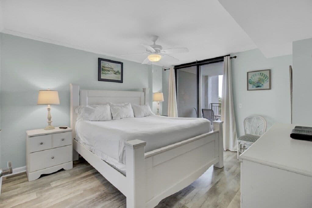 A bedroom with a bed, nightstands, a desk, a chair, a ceiling fan, and a sliding door.