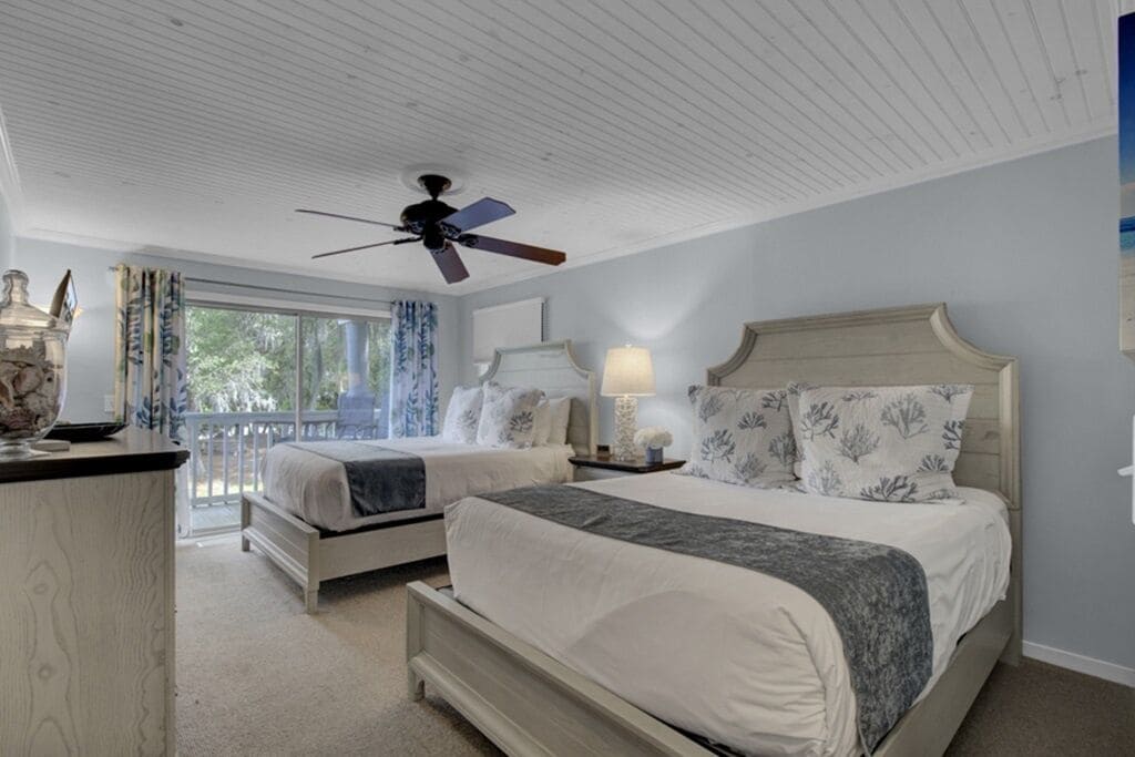 A bedroom with two beds, a ceiling fan, a nightstand with a lamp, and a view of the outdoors.