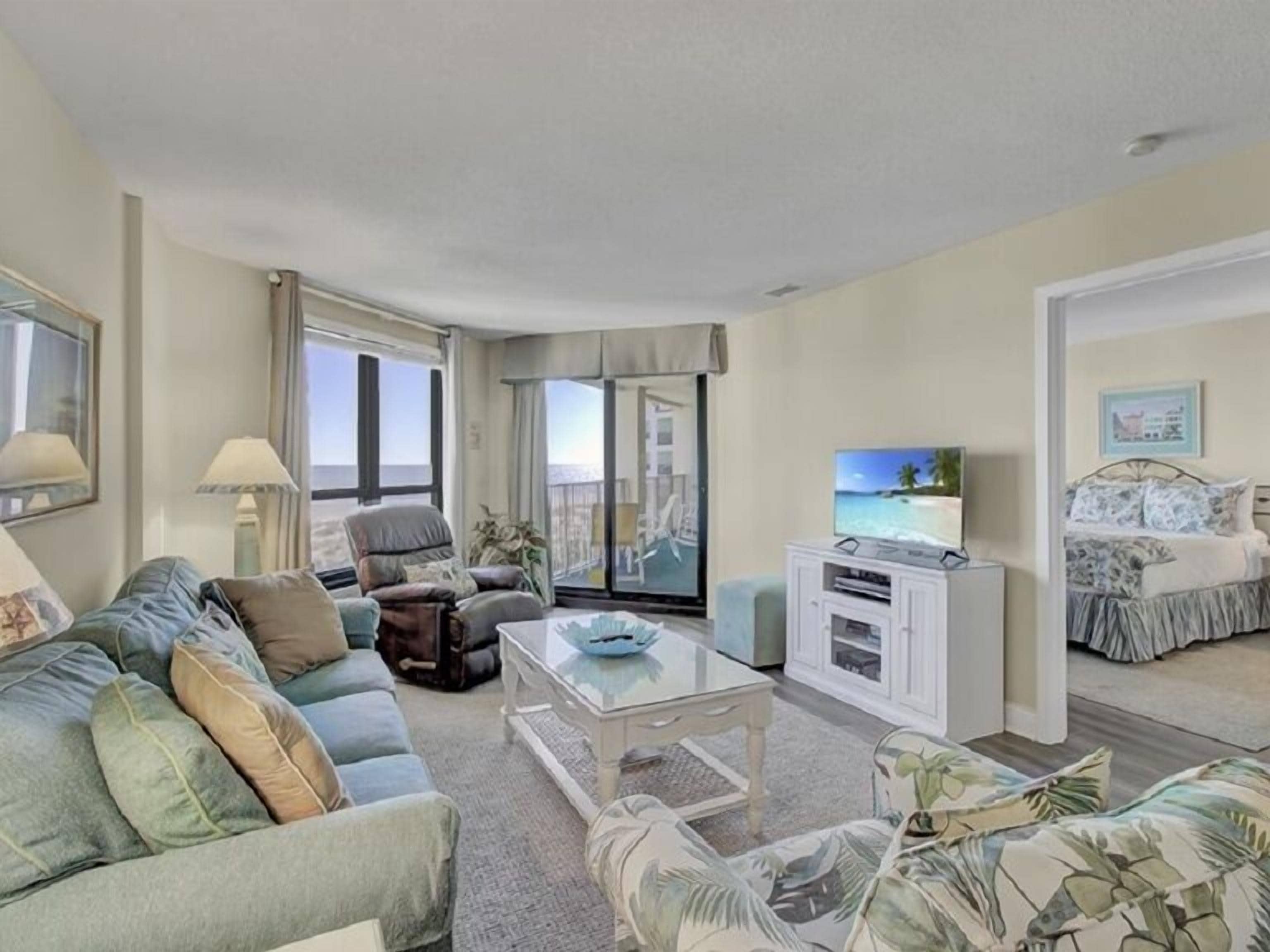 A hotel room with a sofa, armchair, TV, and a bed with a view of the beach.