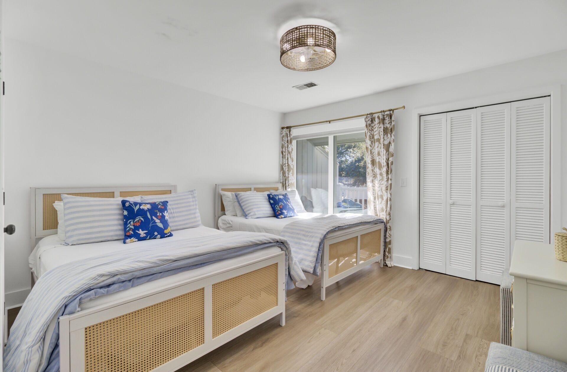 A bedroom with two beds, a window with curtains, a door with louvered shutters, and a ceiling light fixture.