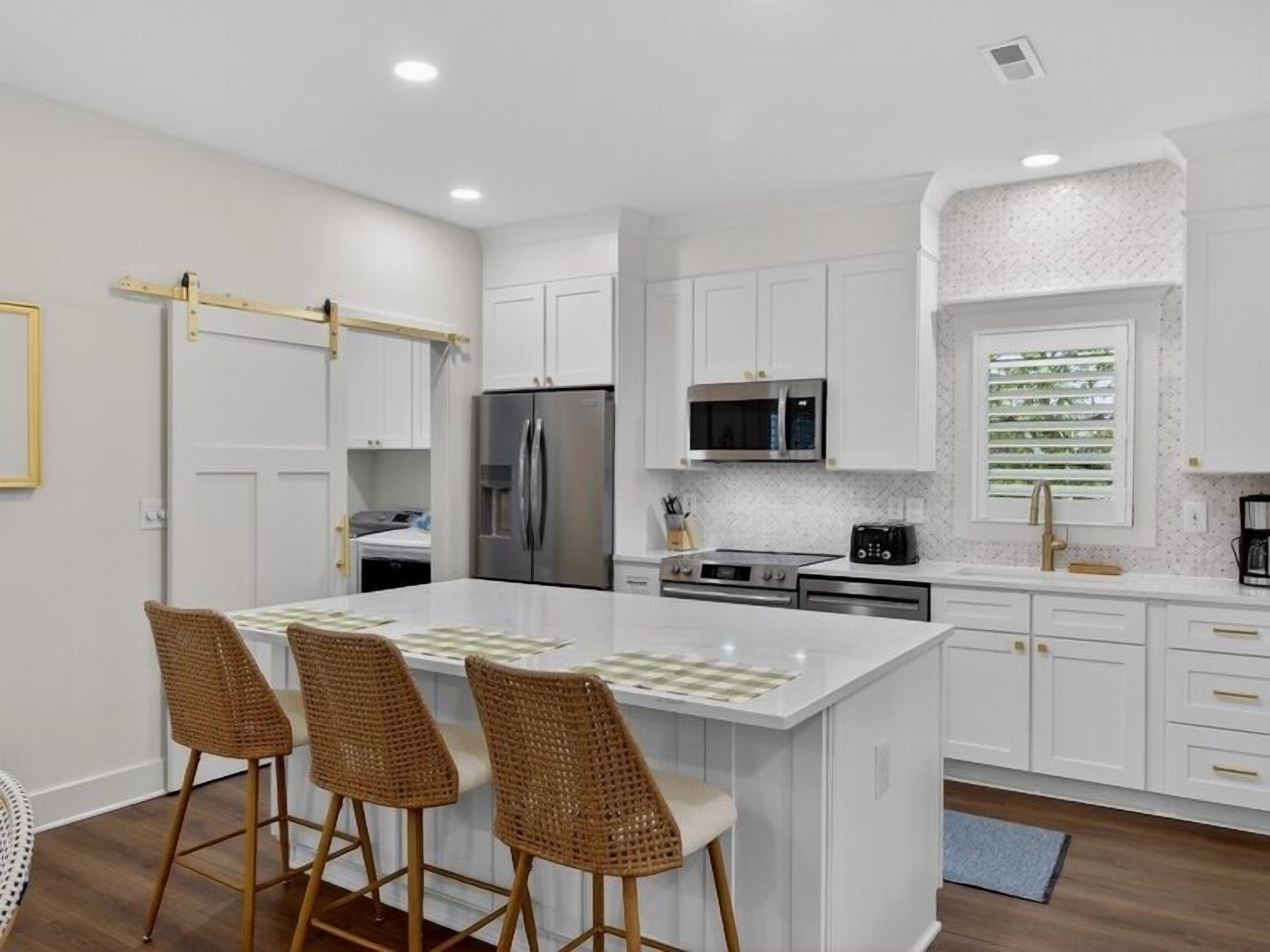 A modern kitchen with white cabinetry, a central island with bar stools, stainless steel appliances, and a window with blinds.
