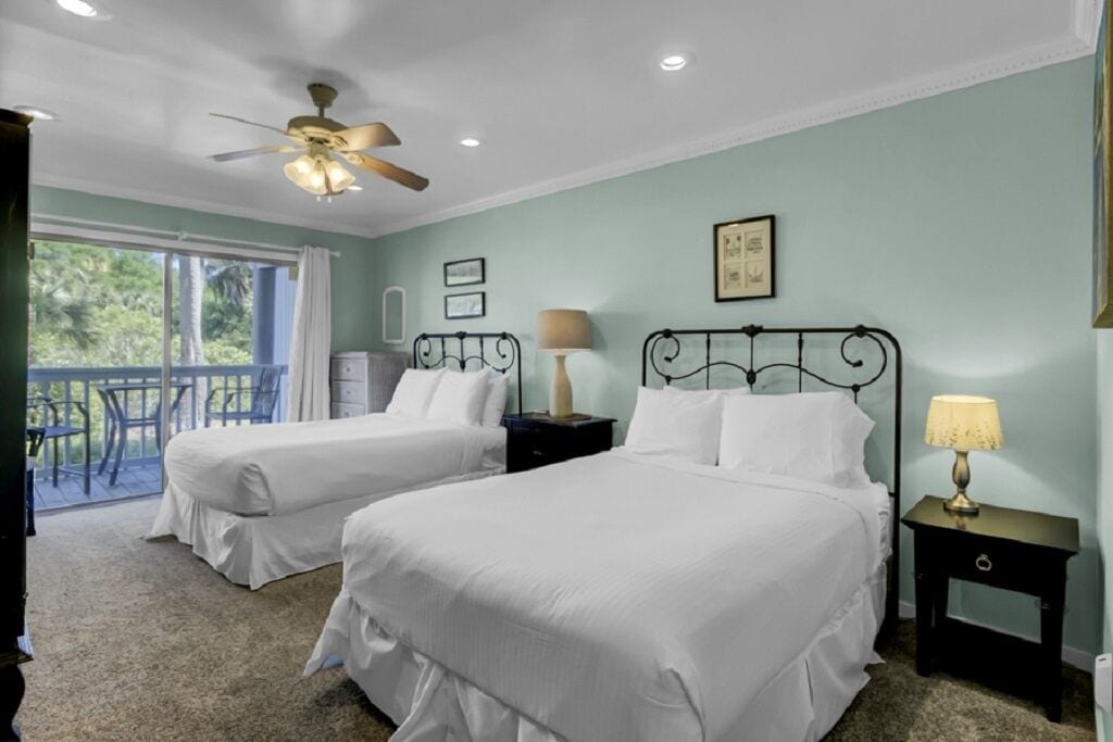 A room with two beds, a ceiling fan, a nightstand with a lamp, and a balcony with outdoor furniture.