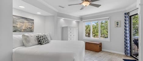 Un dormitorio con cama, ventilador de techo, ventana, cómoda y una pequeña mesita de noche.