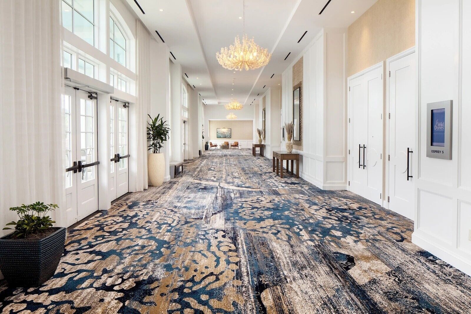 A long hallway with a patterned carpet, white doors, and a chandelier.