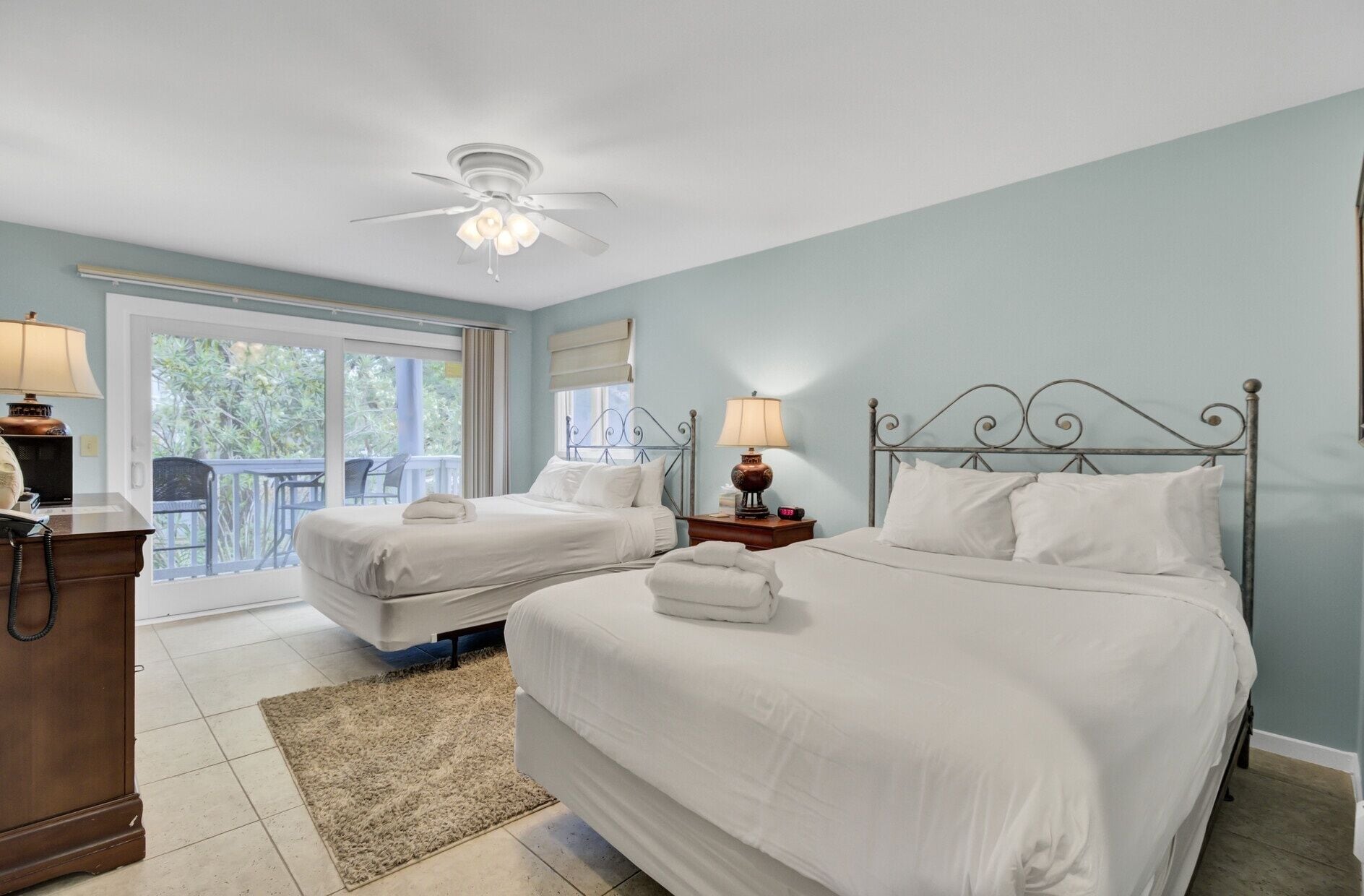 A hotel room with two beds, a ceiling fan, a desk with a lamp, and a view of the outdoors.