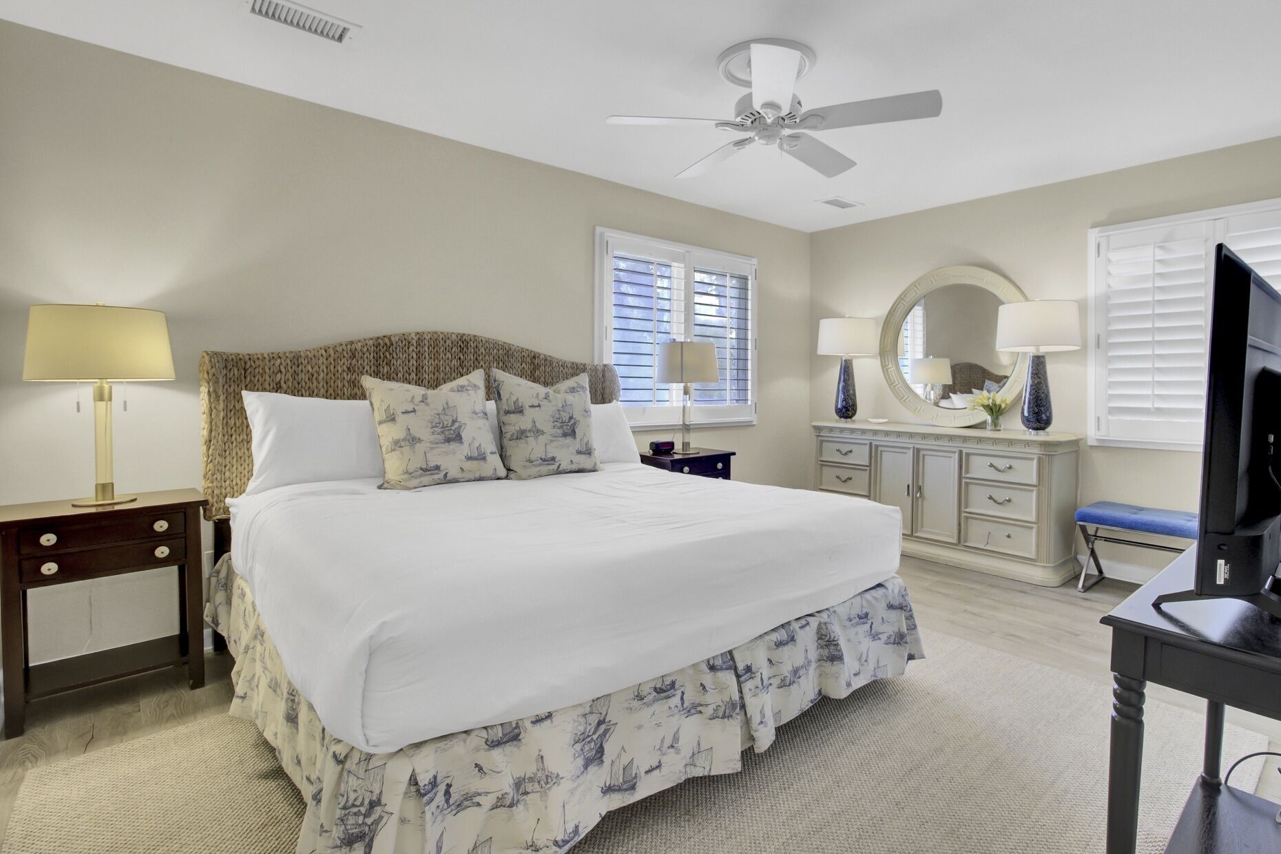 A bedroom with a large bed, a ceiling fan, a dresser, a television, and a window with blinds.