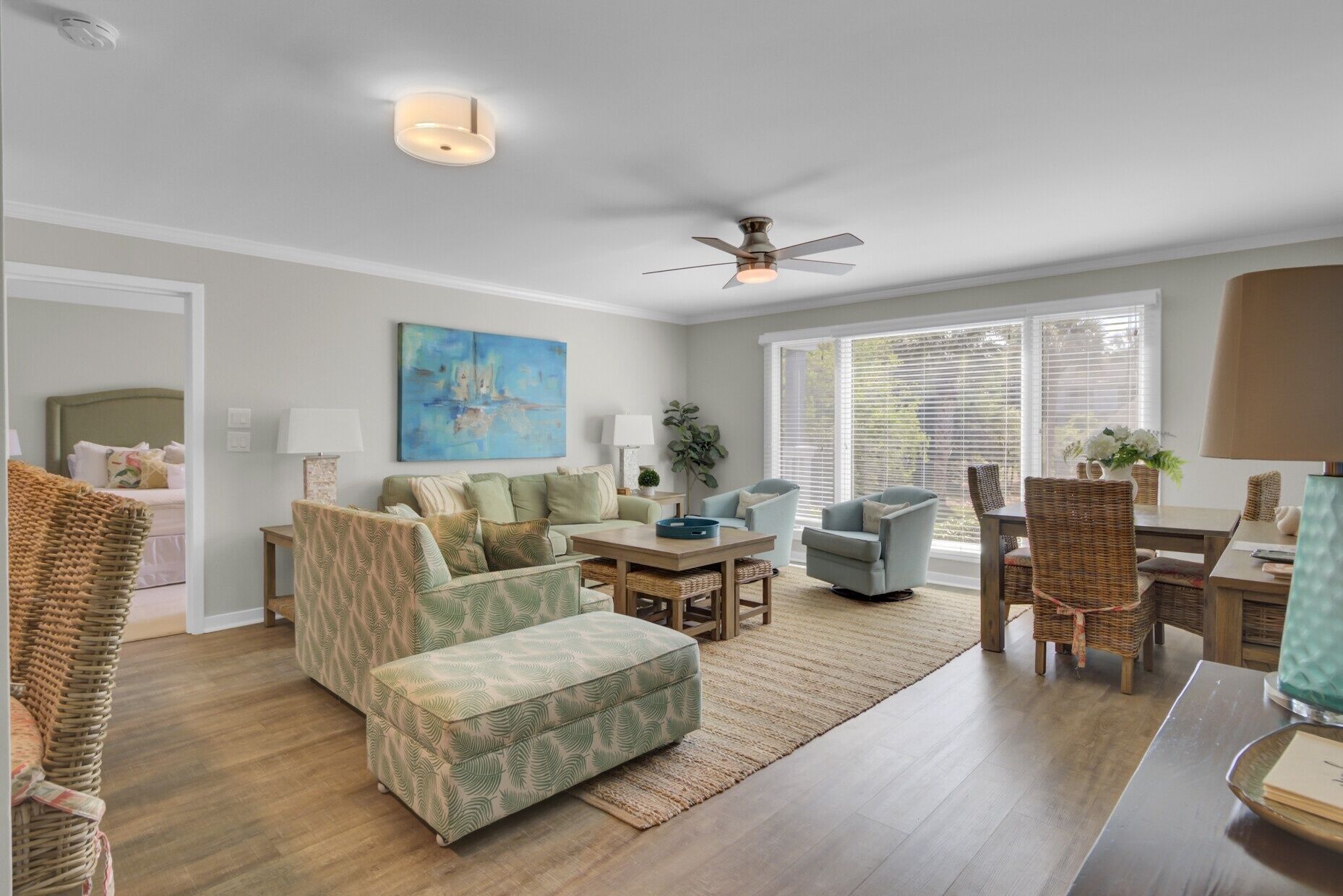 A living room with a sofa, ottoman, and chairs, a dining table, a ceiling fan, and a painting on the wall.