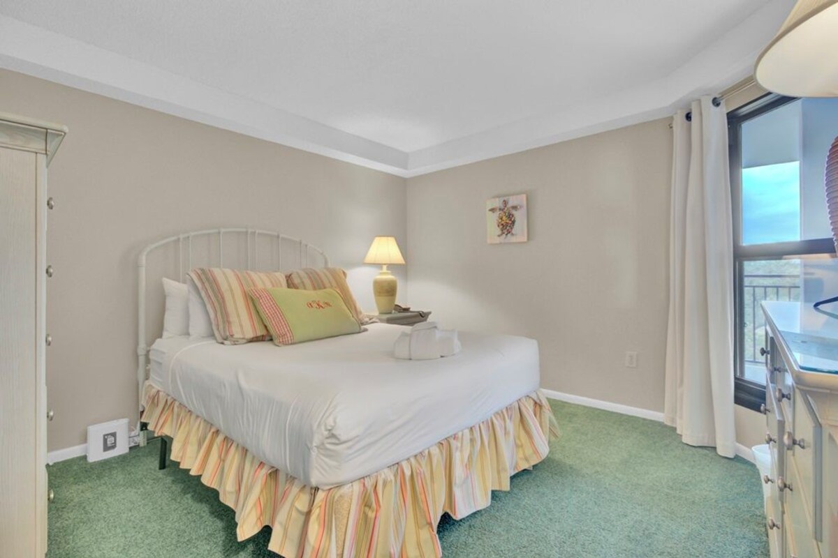 A bedroom with a bed, a bedside lamp, a window with curtains, and a dresser.