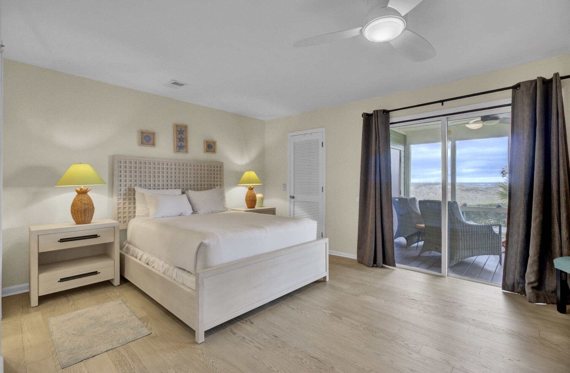 A bedroom with a bed, two bedside tables with lamps, a ceiling fan, a sliding glass door to a balcony, and a view of the landscape.