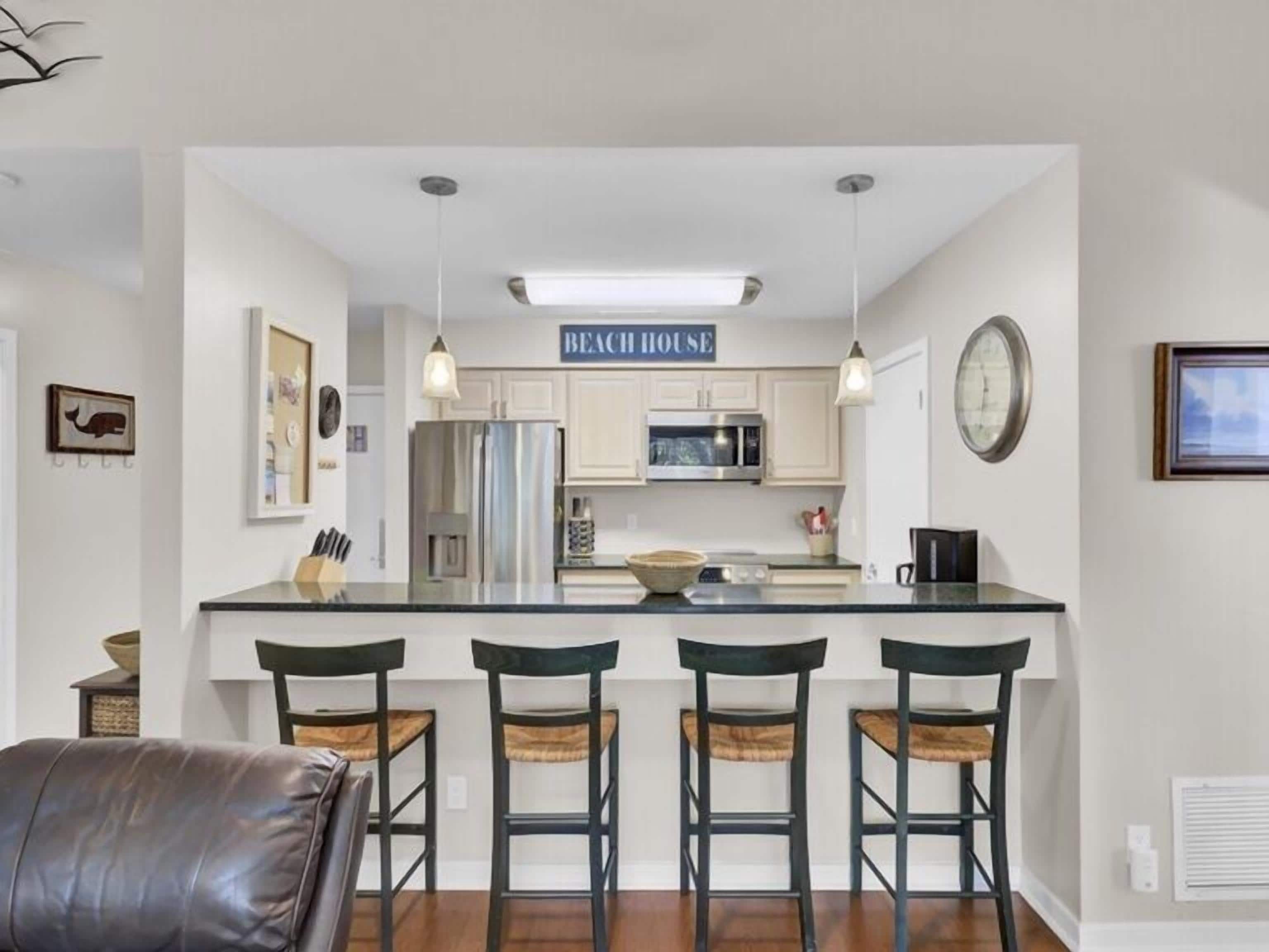 A kitchen with a central island, bar stools, a refrigerator, microwave, and a sign that reads 'BEACH HOUSE'.