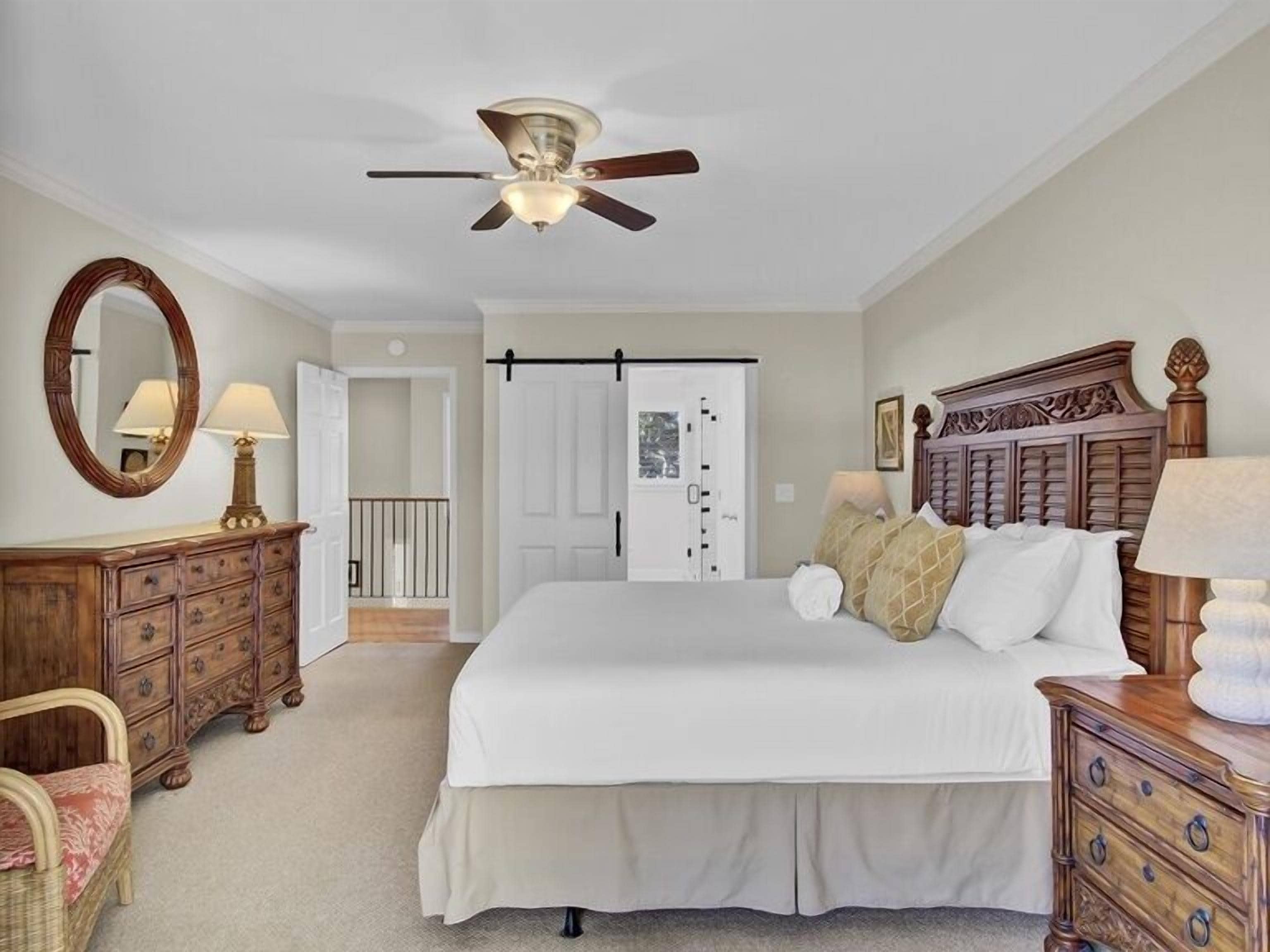 A bedroom with a bed, wooden dresser, chair, ceiling fan, and a door leading to another room.