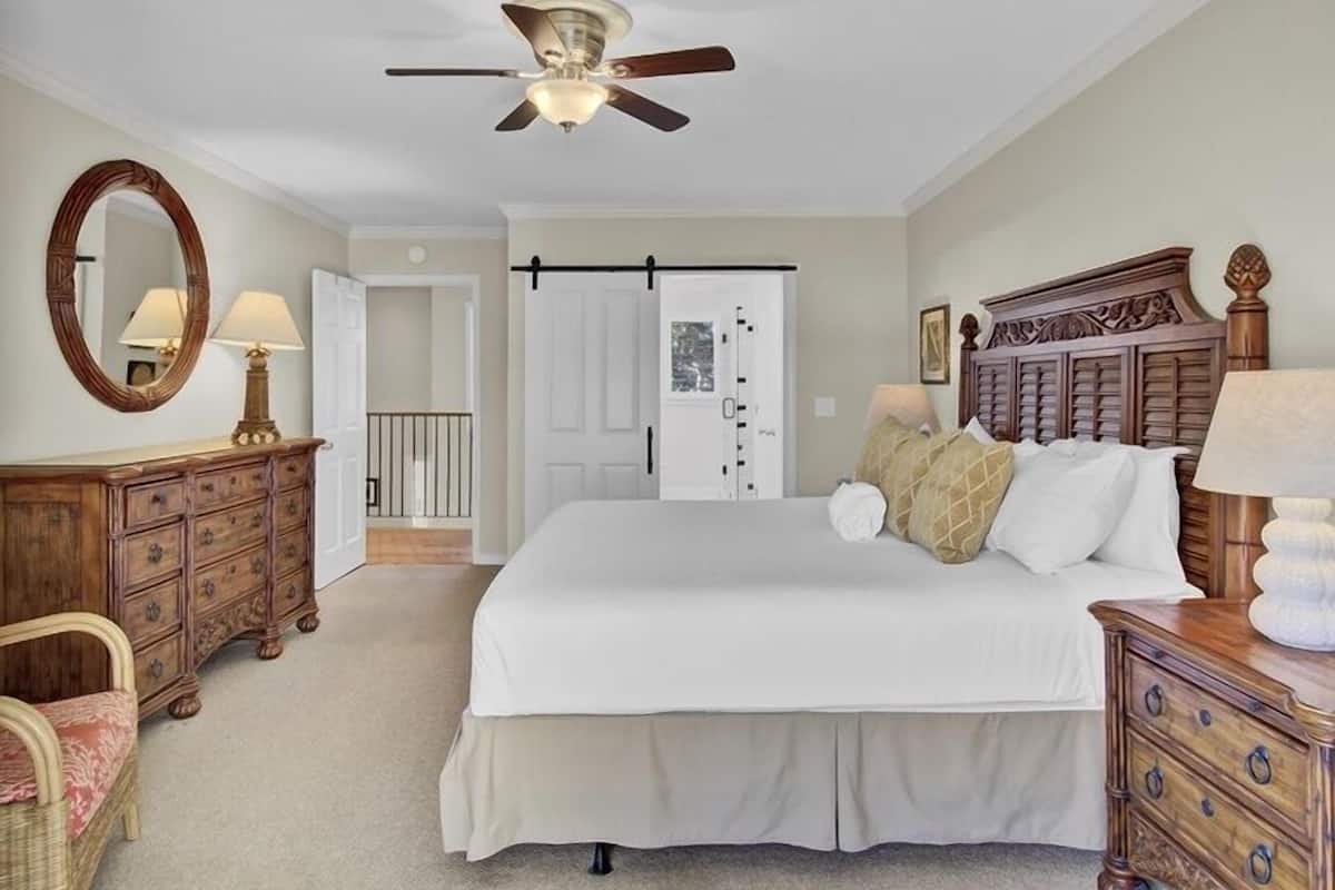 Un dormitorio con una cama, cómoda de madera, una silla, ventilador de techo y una puerta que da a otra habitación.