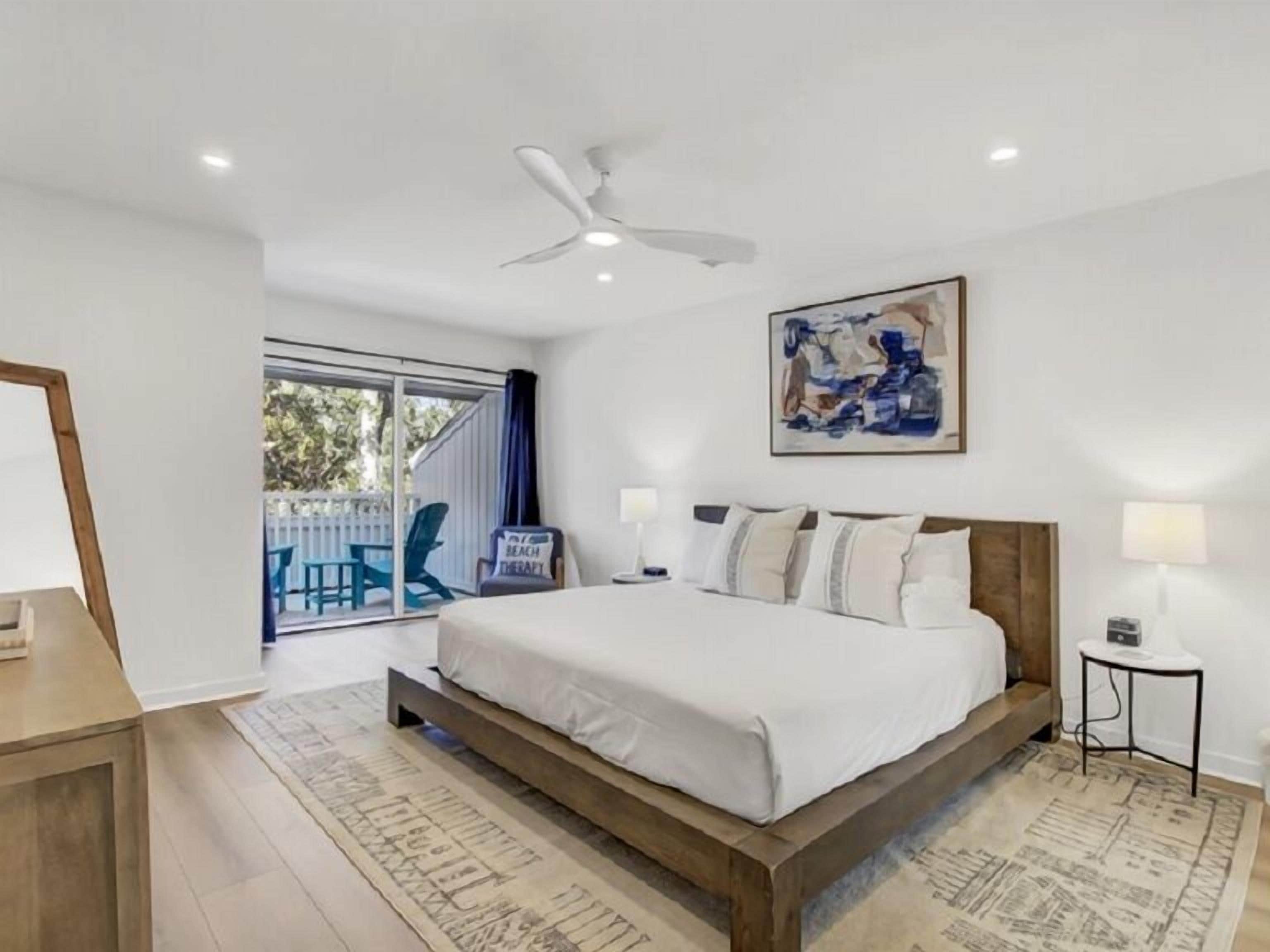 A bedroom with a large bed, a ceiling fan, a painting, and a view of the outside.