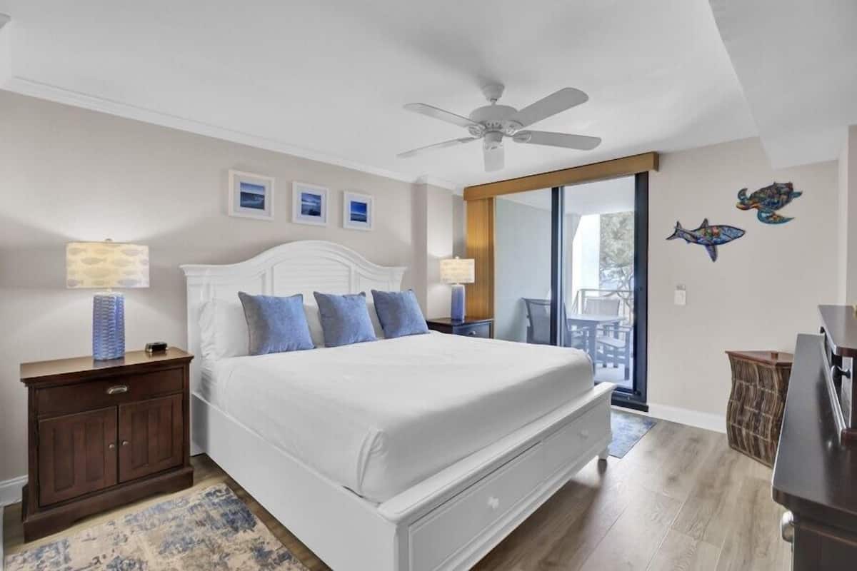 Un dormitorio con cama, mesitas de noche, ventilador de techo, ventana grande y vistas al exterior.