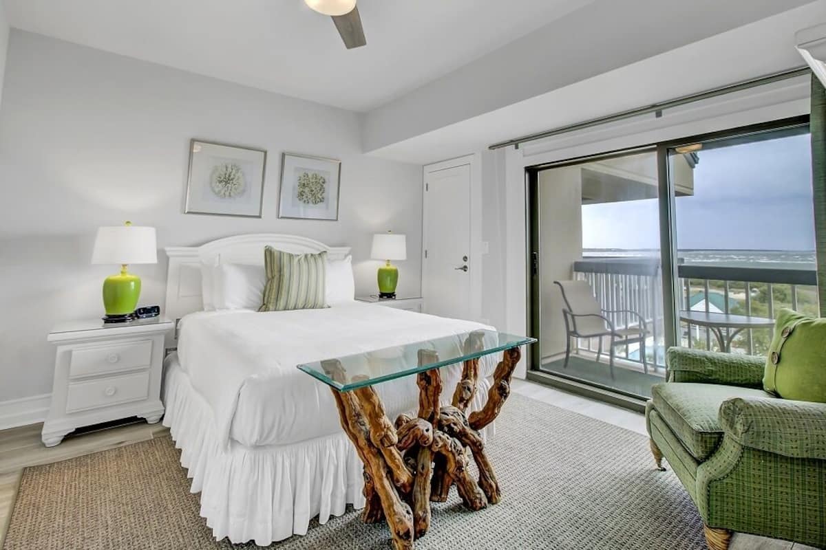 Un dormitorio con cama, mesitas de noche, una mesa de centro de vidrio, una silla verde y un balcón con vistas.
