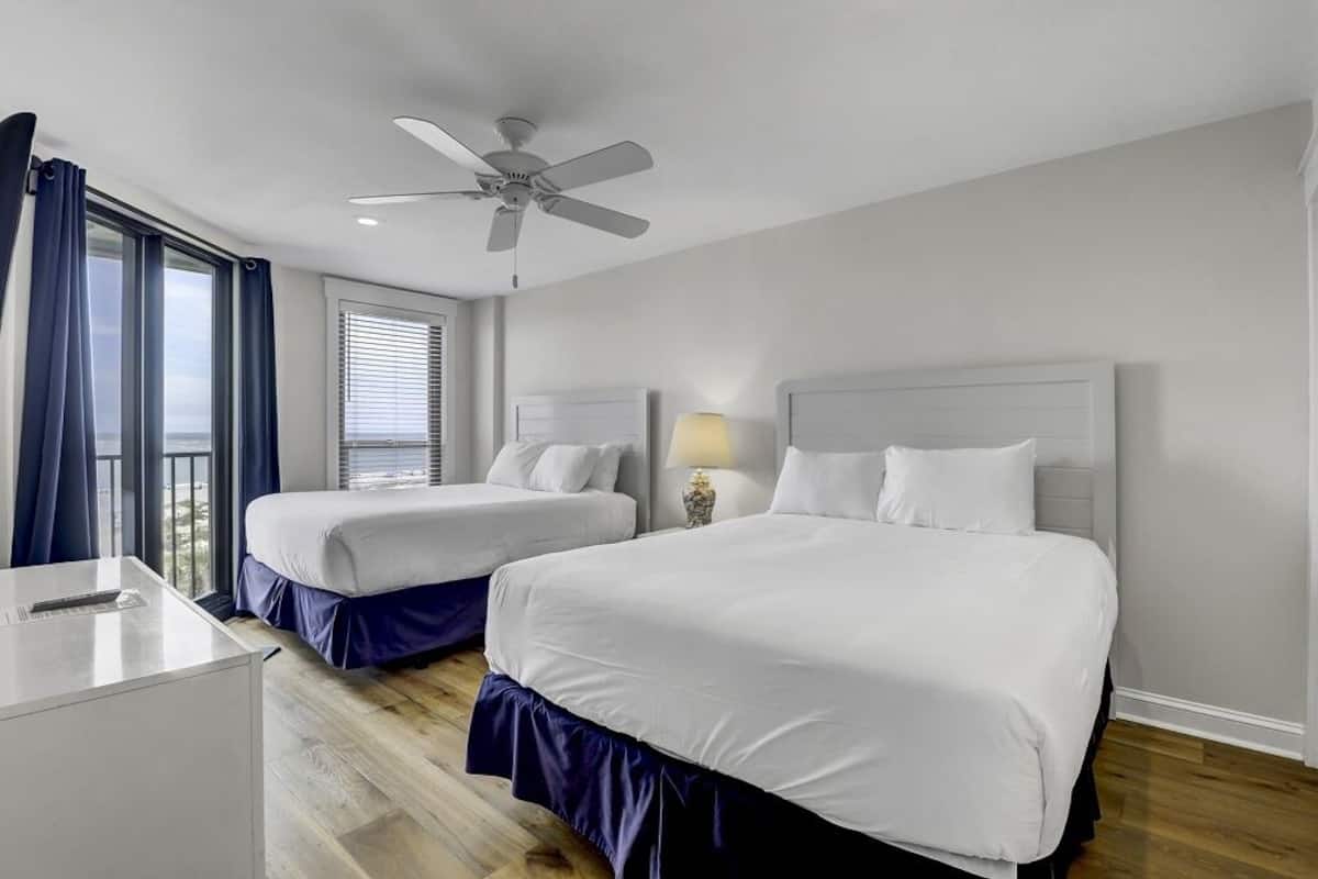 Una habitación de hotel con dos camas, un escritorio, un ventilador de techo y una ventana con cortinas.